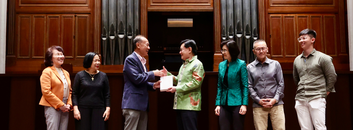 Group of seven people on a stage; two people exchange a gift. Pipe organ background.